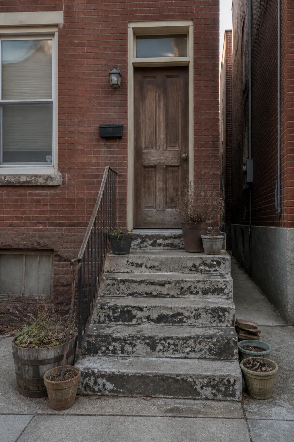 Weathered concrete steps leading to a worn wooden door on red brick rowhouse, potted plants.