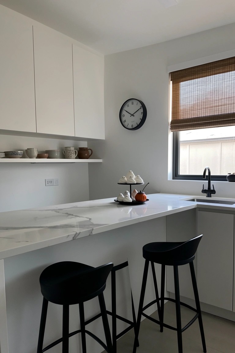 Modern white kitchen with marble counter, black stools, ghost decorations, and wall clock.