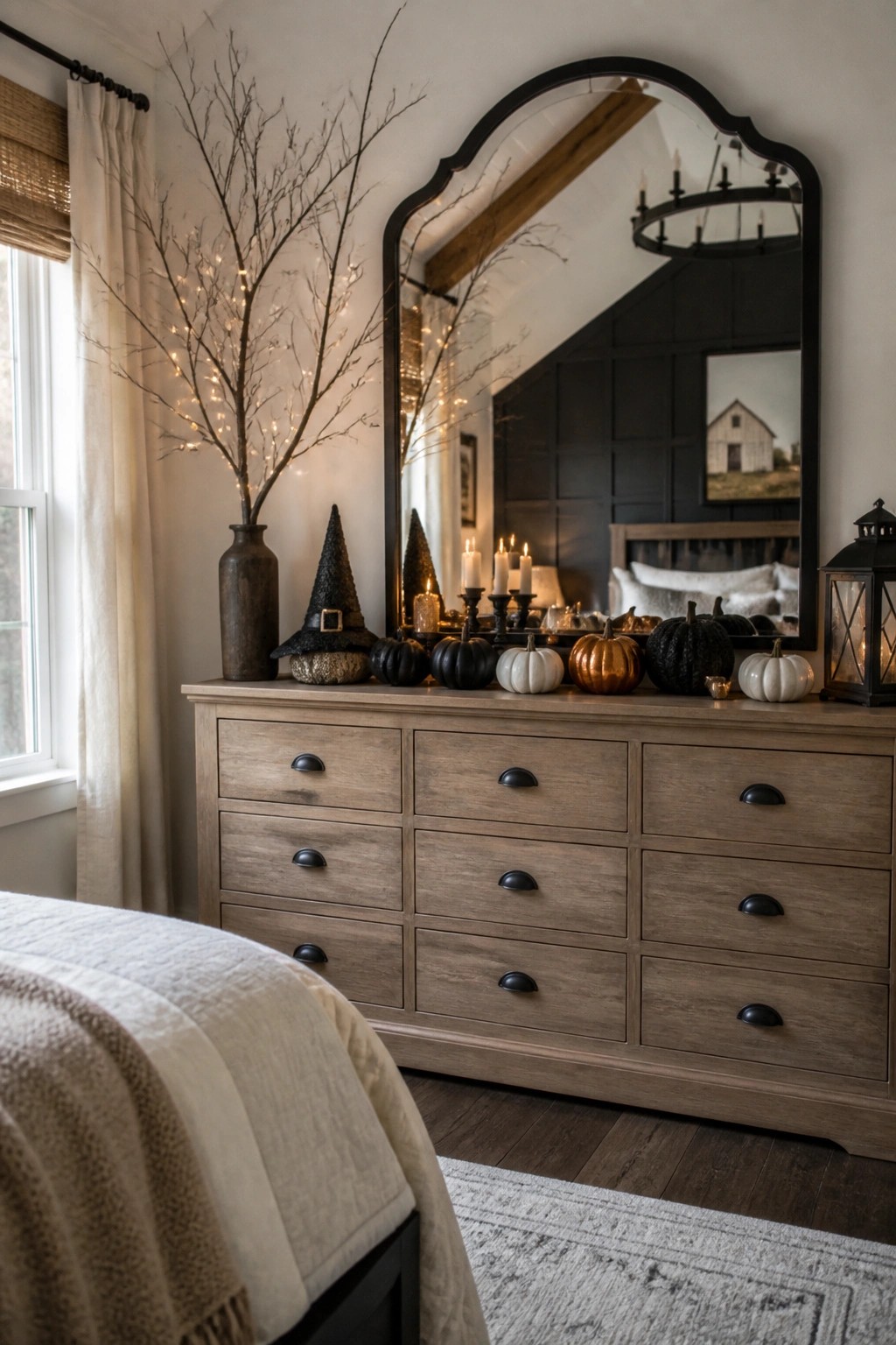 Bedroom dresser with a black witch hat, black white and orange pumpkins, lit branches, and candles arranged beneath a large arched mirror.