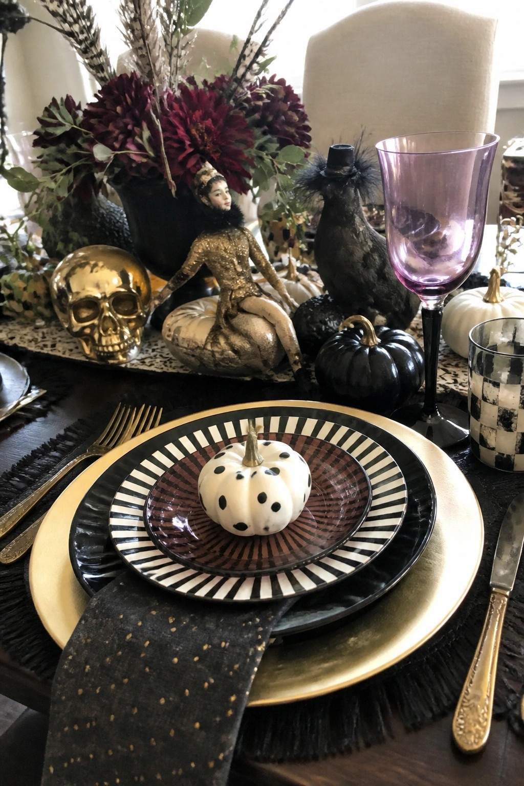 Dining table with stacked black and gold plates holding a small white polka dot pumpkin, placed beside a gold skull and a sequined skeleton figurine.