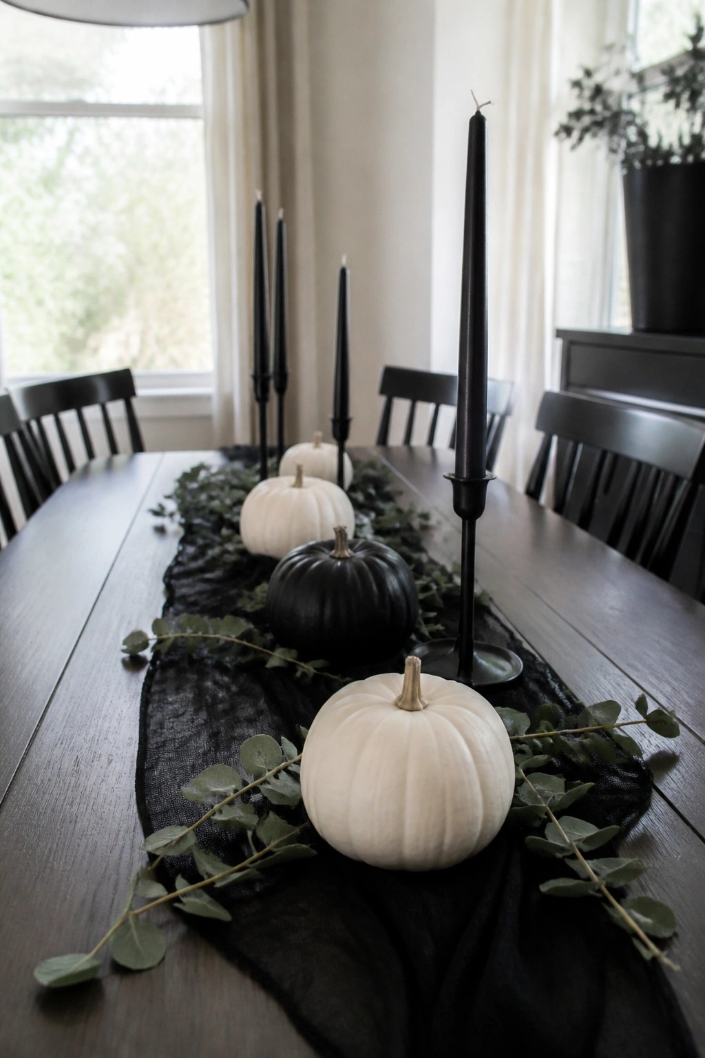 Dining table with a black runner holding white and black pumpkins, tall black candles, and scattered eucalyptus.