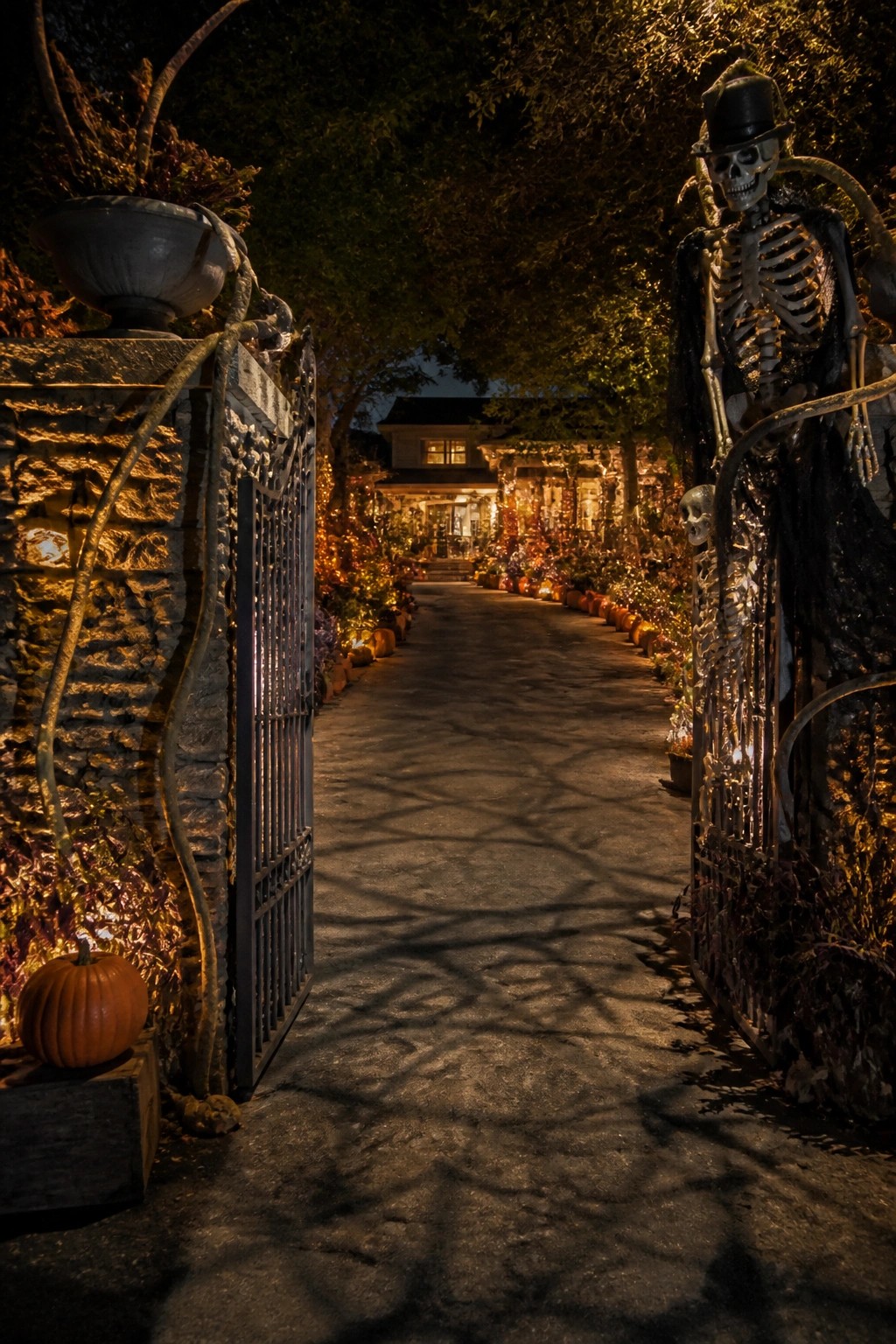 Nighttime view of an outdoor walkway lined with pumpkins and string lights, with large skeleton figures positioned beside open metal gates.