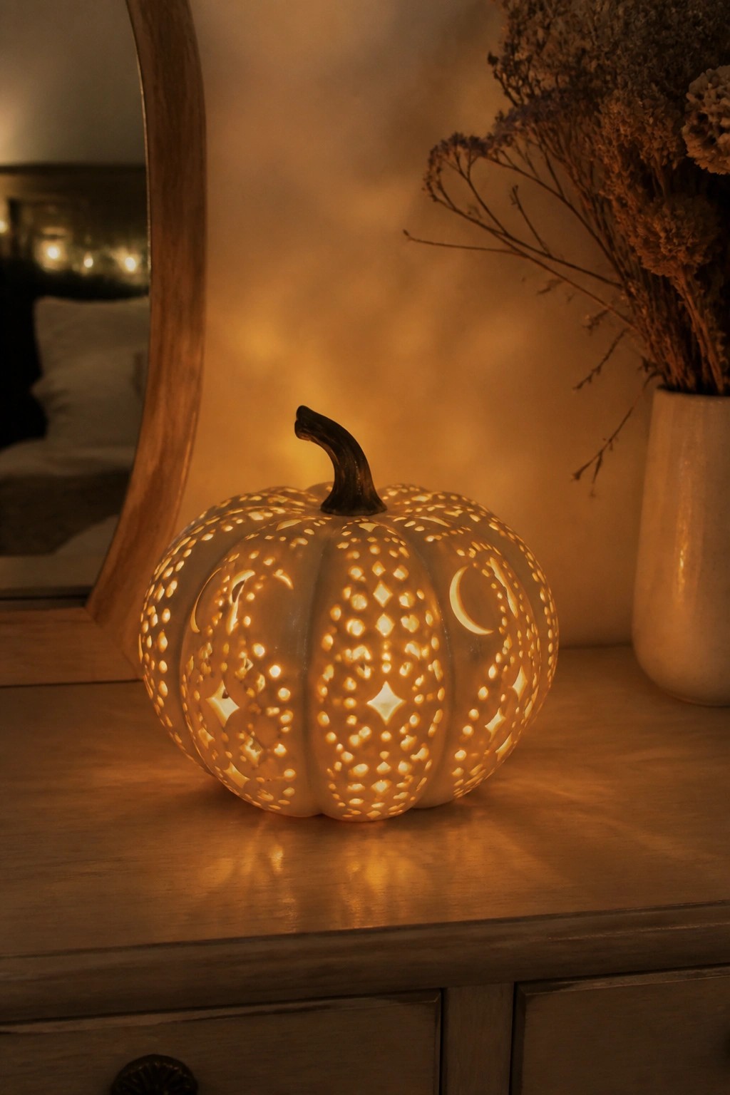A white pumpkin carved with moons, stars, and diamond patterns glowing on a wooden dresser beside a vase of dried flowers.
