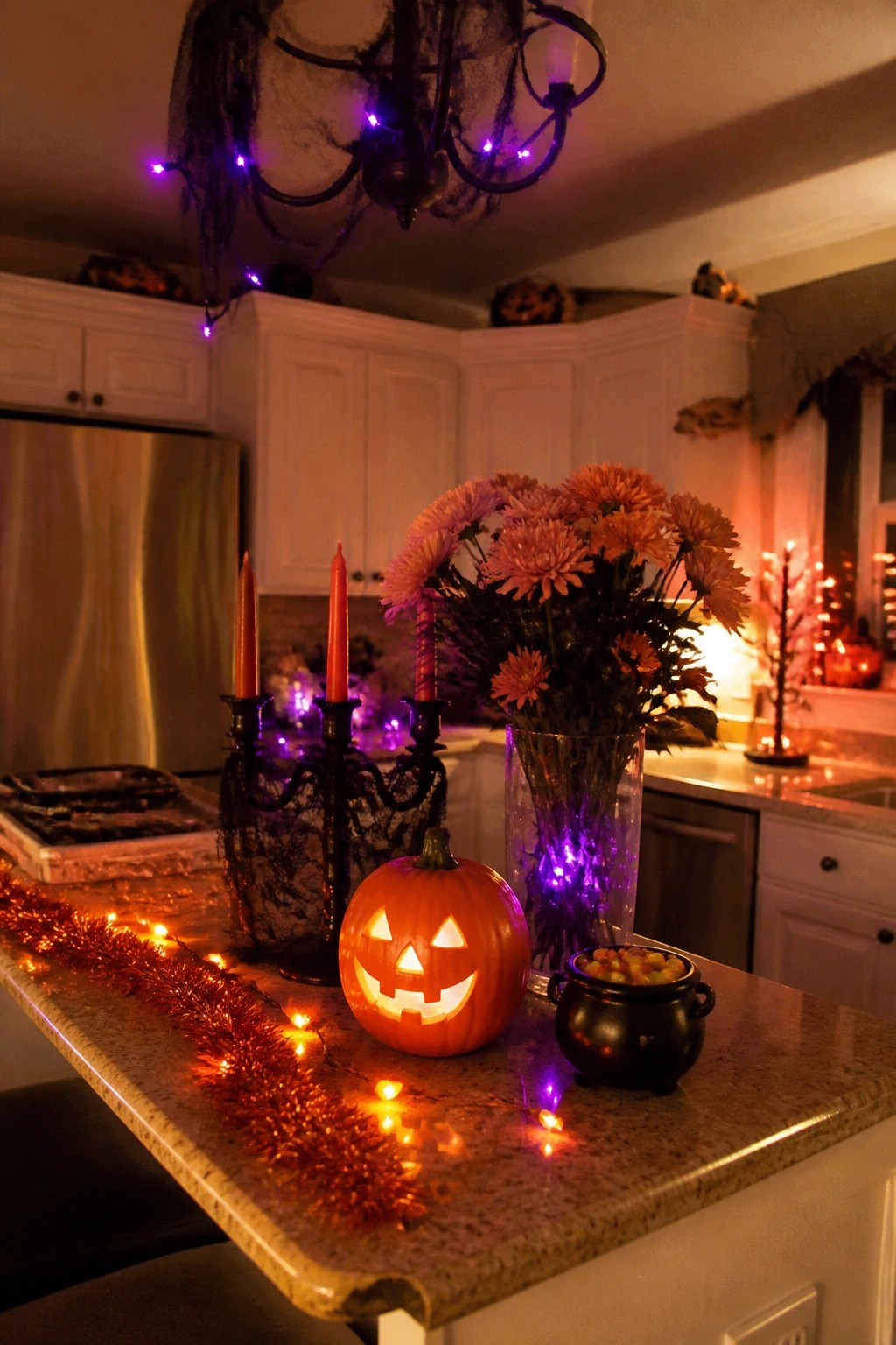 A lit jack-o'-lantern sits on a kitchen island with orange string lights, red candles, and a black cauldron nearby, while spiderwebs and purple lights decorate the chandelier above.