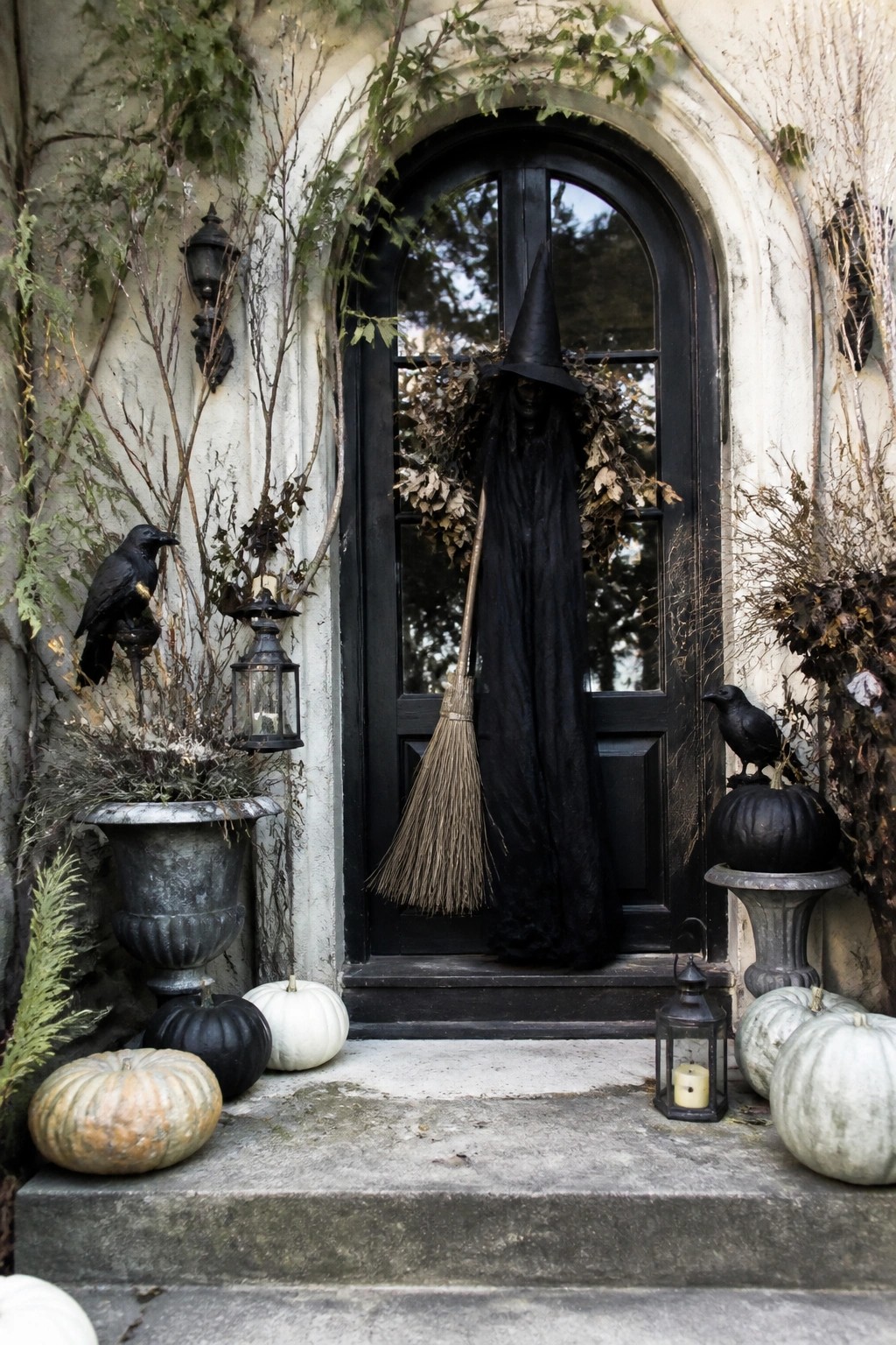 Front door with a tall witch figure in a black cloak and pointed hat holding a broom, flanked by black crows and pumpkins on the steps.