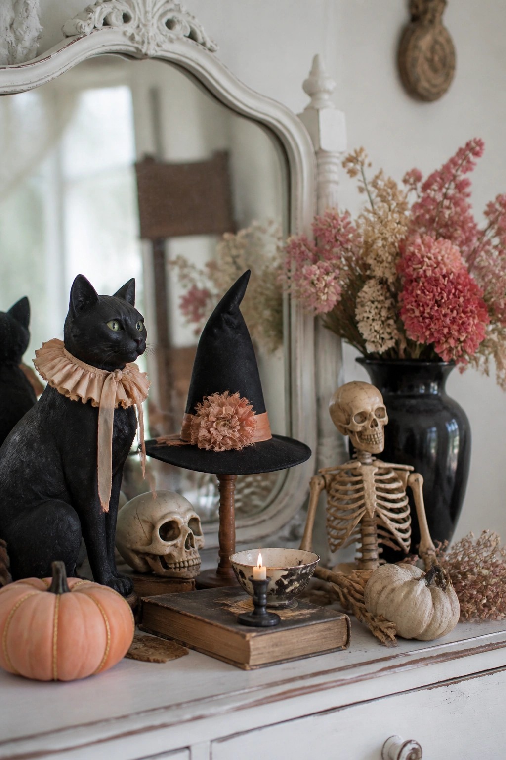 A black cat figurine with a ruffled collar sits beside a black witch hat on a white console, with skulls, a small skeleton, pumpkins, and dried flowers in the background.