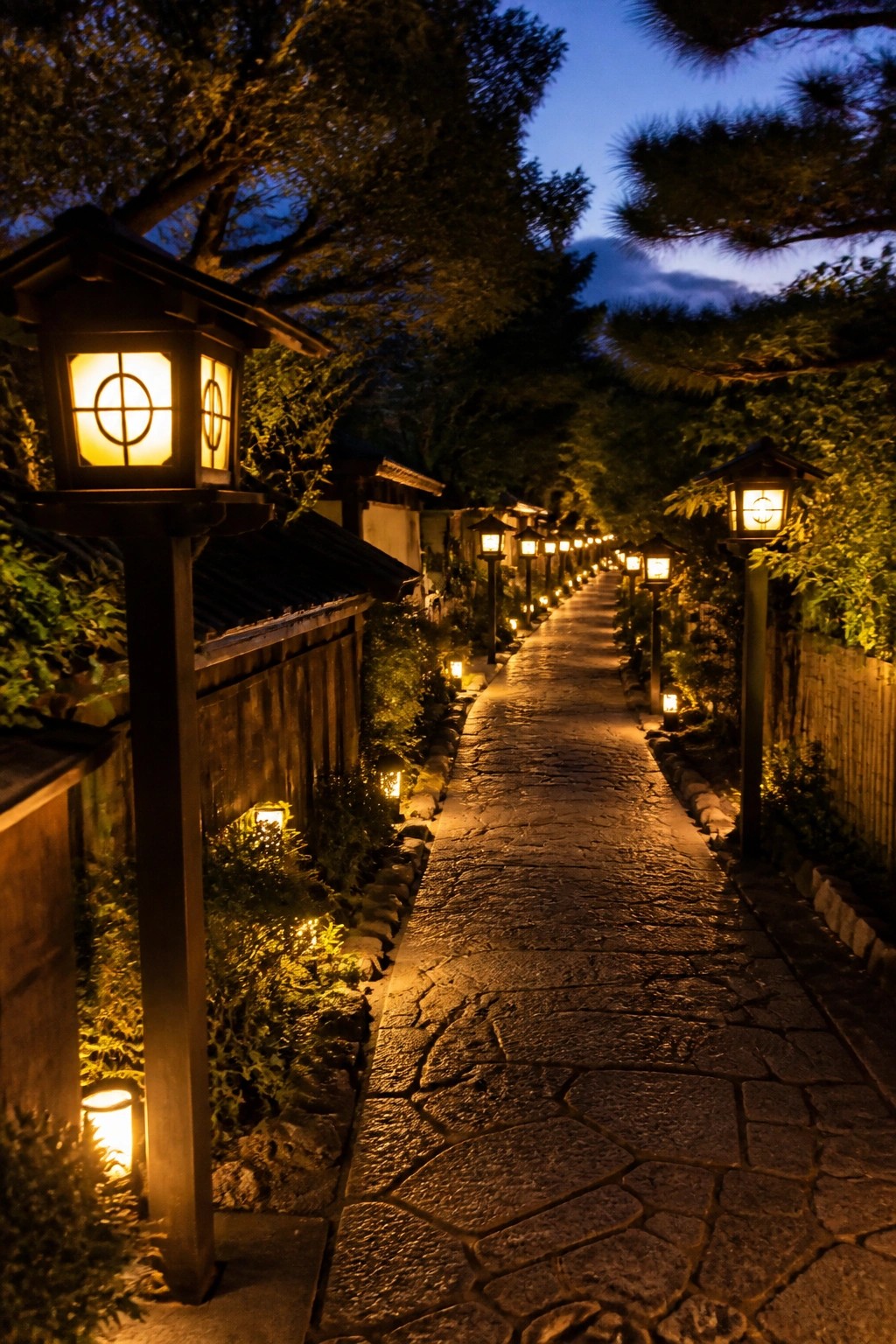 Stone walkway lined with traditional lanterns on posts glowing at dusk in a garden setting.