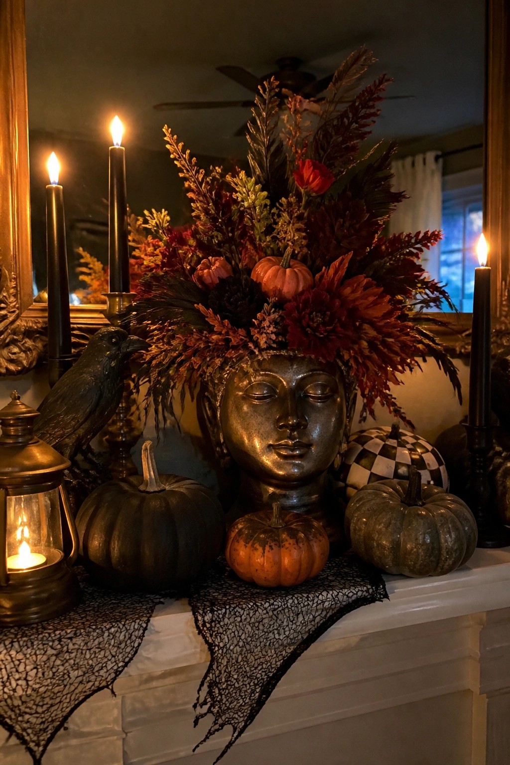 A white fireplace mantel decorated for Halloween with a bronze Buddha head centerpiece topped with dark florals and small pumpkins, black taper candles, a crow figurine, and assorted pumpkins arranged on a black lace runner.