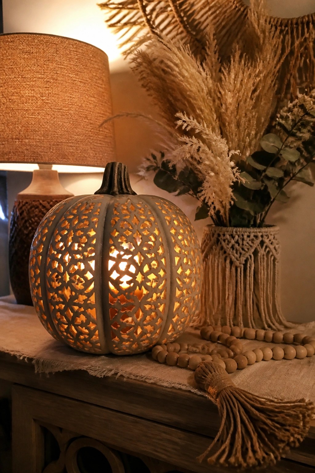 A carved gray pumpkin lantern with intricate cutouts glowing from within, placed on a wooden console beside a table lamp and dried grasses in a macrame vase.