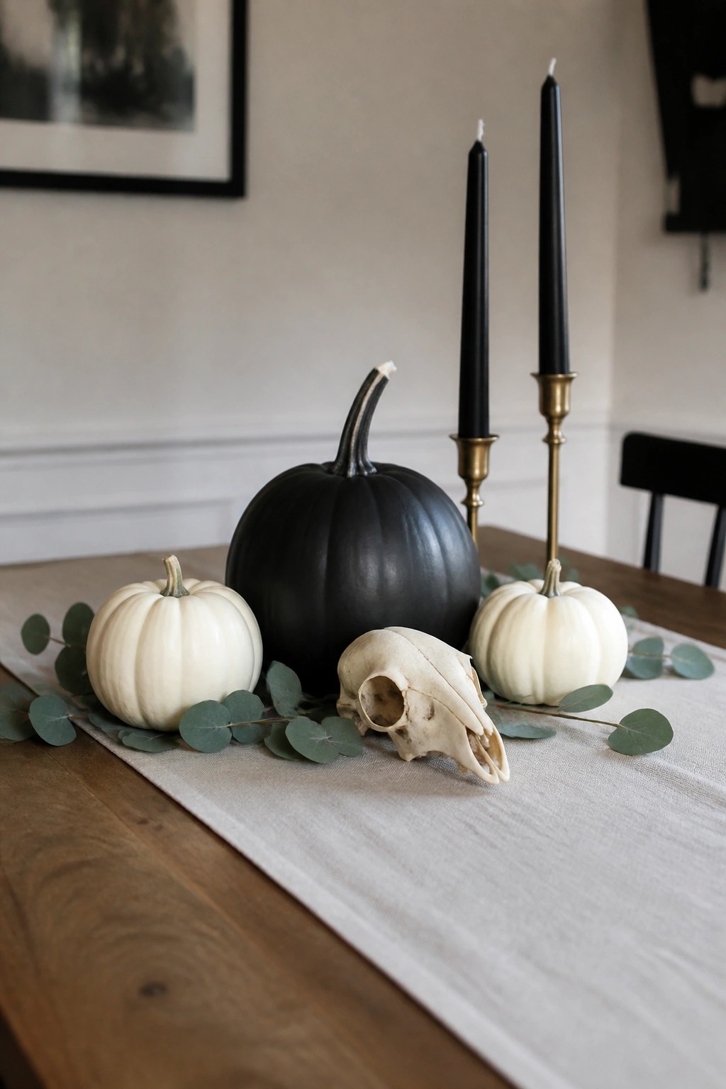 Dining table with a central animal skull, black and white pumpkins, black taper candles, and eucalyptus on a linen runner.