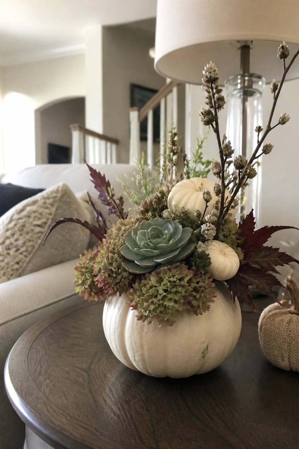 White pumpkin vase with succulent, dried flowers, red leaves on wooden table beside lamp