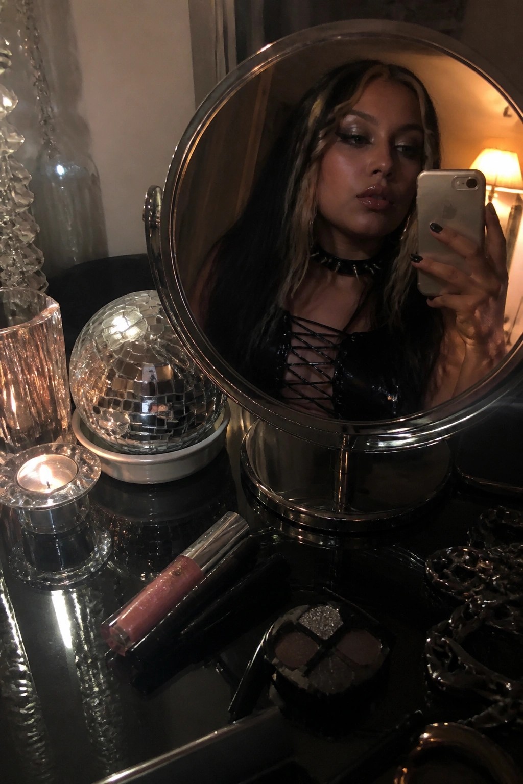 Round vanity mirror on a dark table reflecting a woman taking a selfie, with a disco ball, lit candle, and scattered makeup products in front of it.