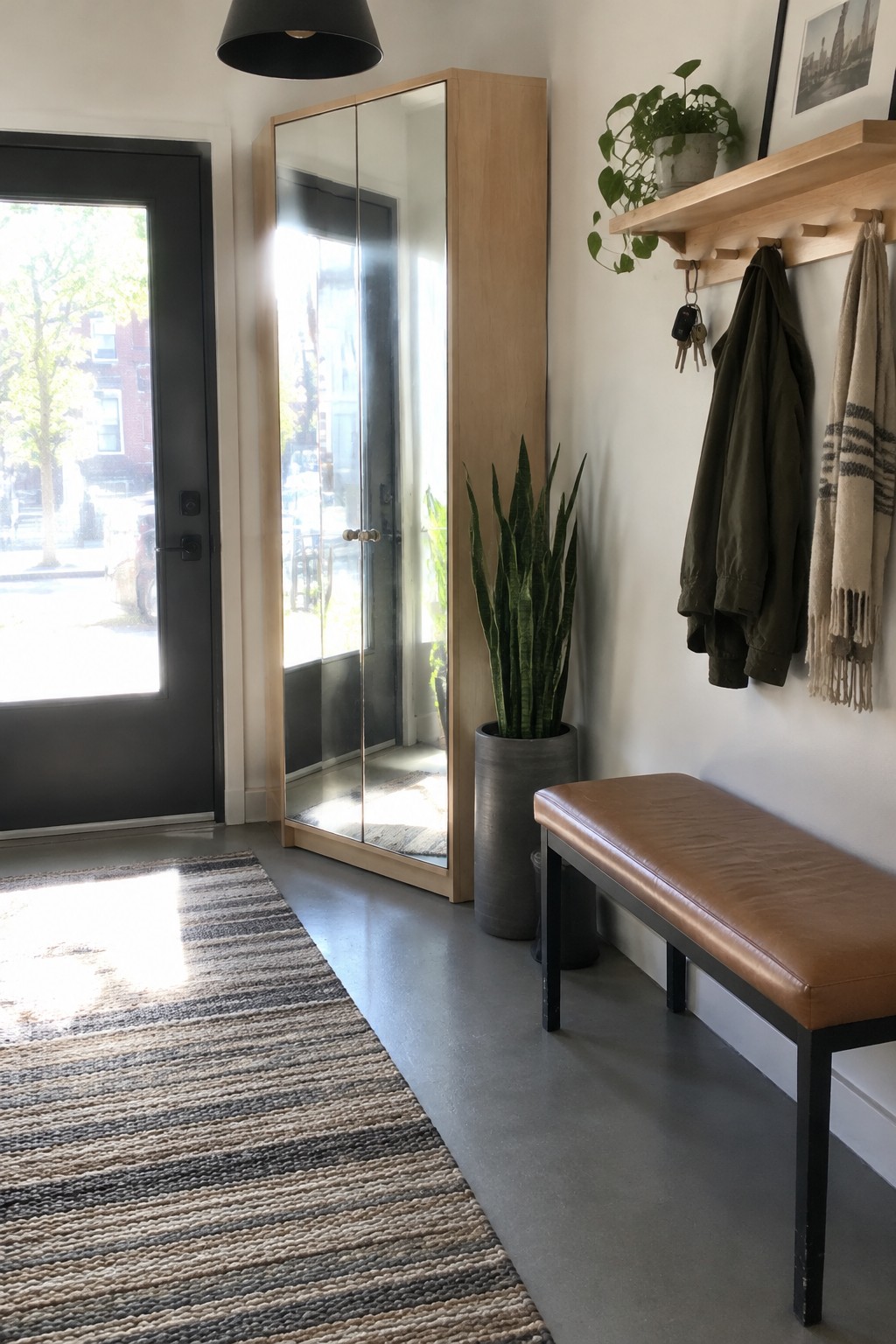 A mirrored wood cabinet stands beside a dark front door in an entryway with a bench, coat hooks, and a striped rug.