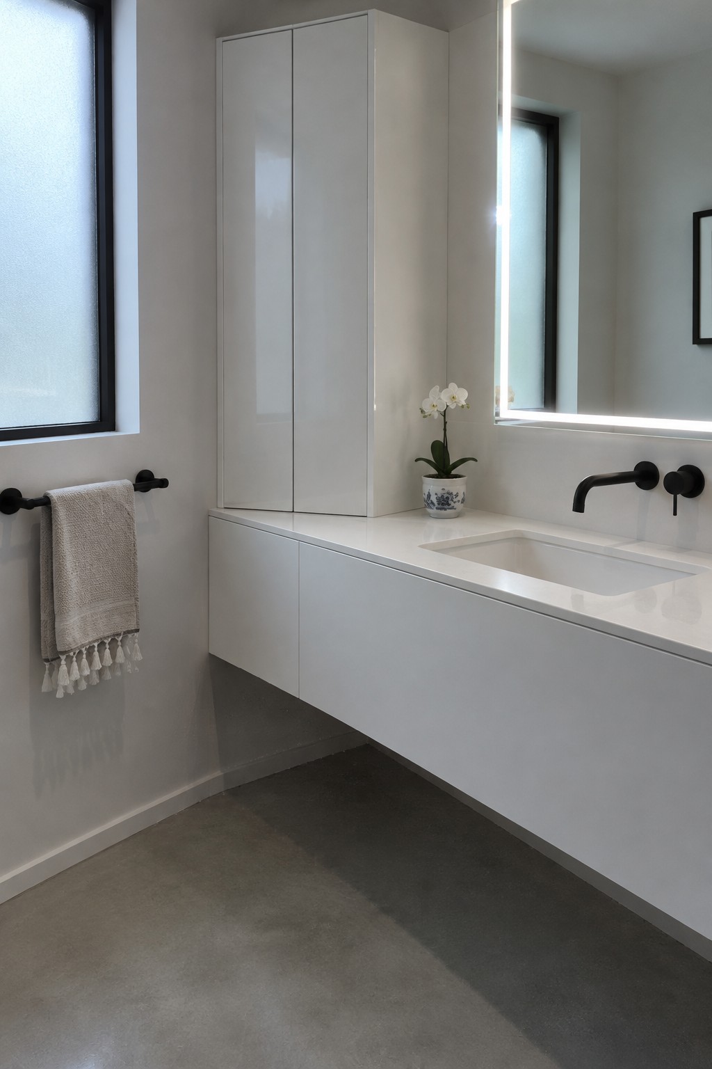 A white bathroom with a tall corner cabinet, floating vanity, rectangular sink, and black wall-mounted faucet.