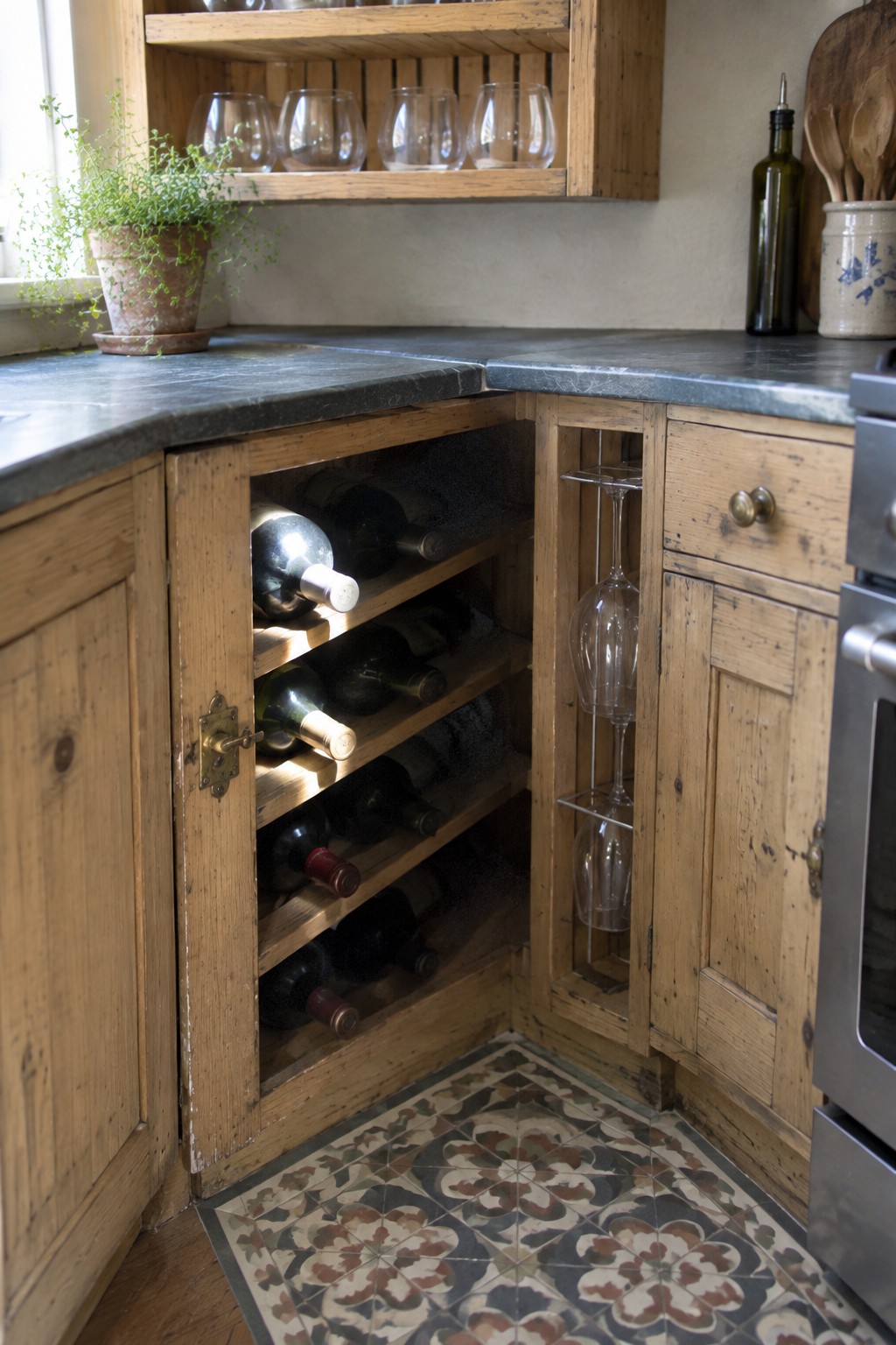 An open wooden corner cabinet holds wine bottles on angled shelves beside a narrow rack for wine glasses.