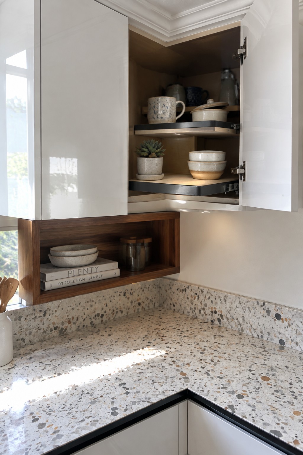 An open white corner cabinet with dishes and a small potted plant on two shelves above a terrazzo countertop.